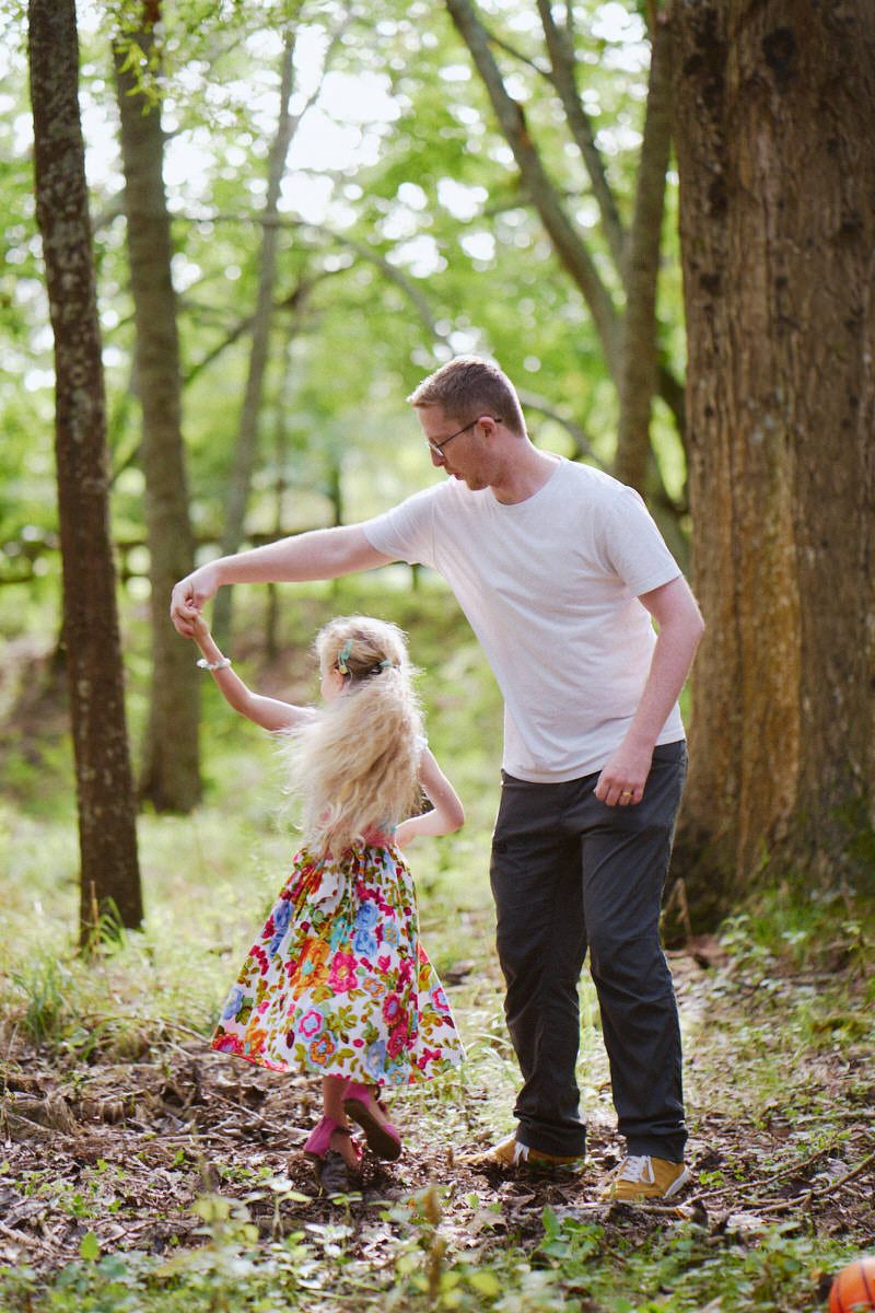 Father and daughter dancing together in a sunlit forest, girl wearing a colorful floral dress.
