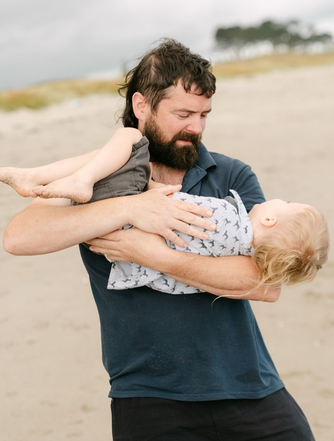 Bearded father playfully swinging his blonde toddler on a sandy beach, both enjoying a joyful moment outdoors.
