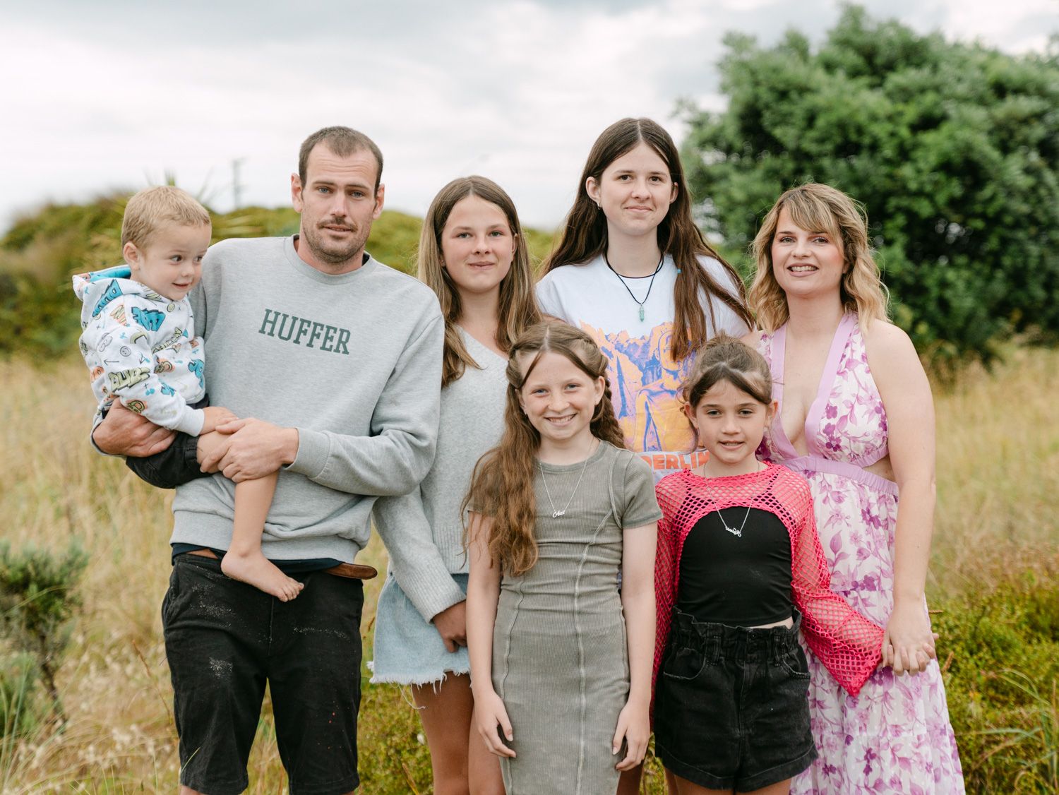 A happy family of seven posing together outdoors in a grassy field on a cloudy day.