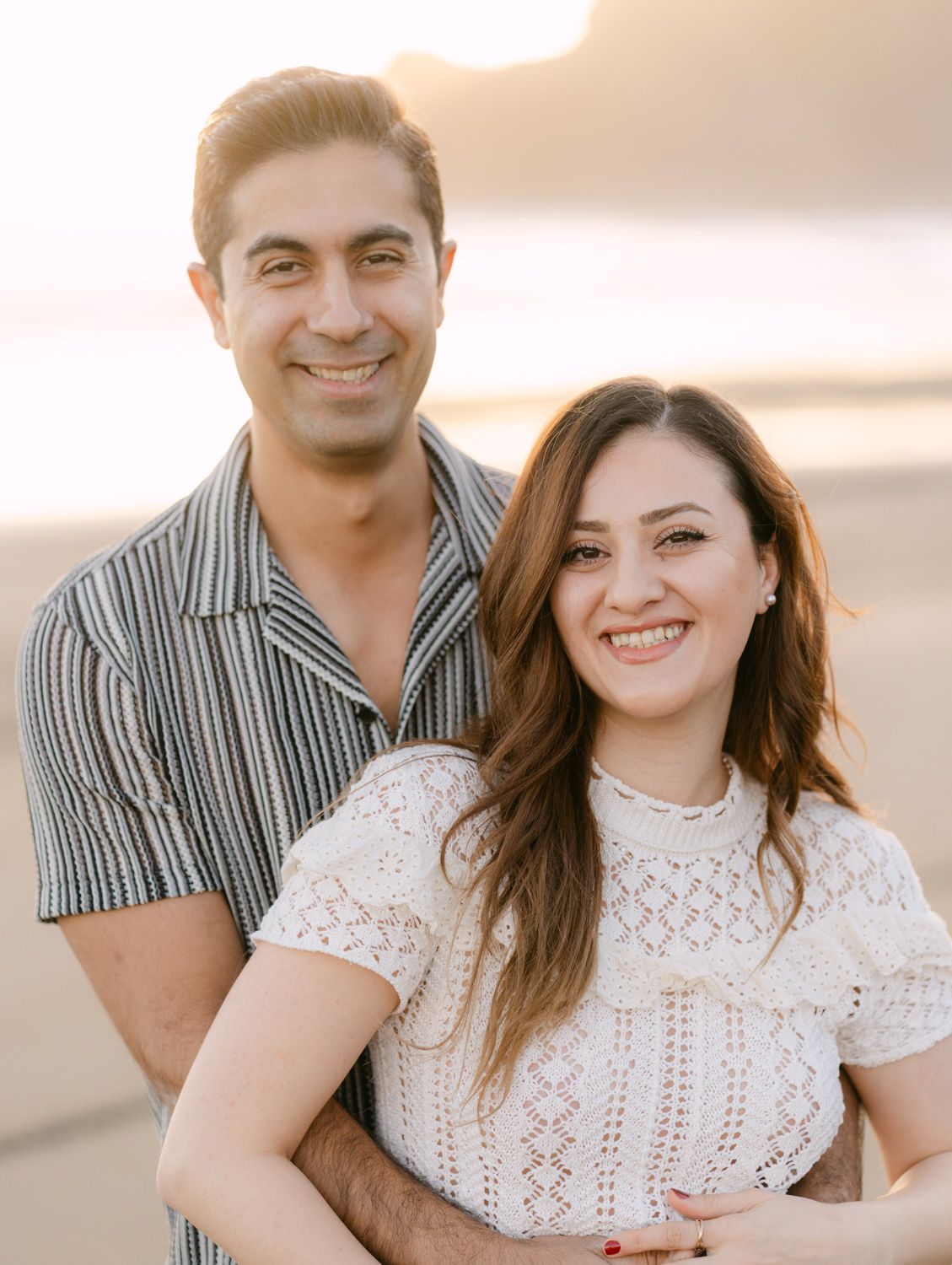 A smiling couple poses together on a beach at sunset, woman in white lace top and man in striped shirt.