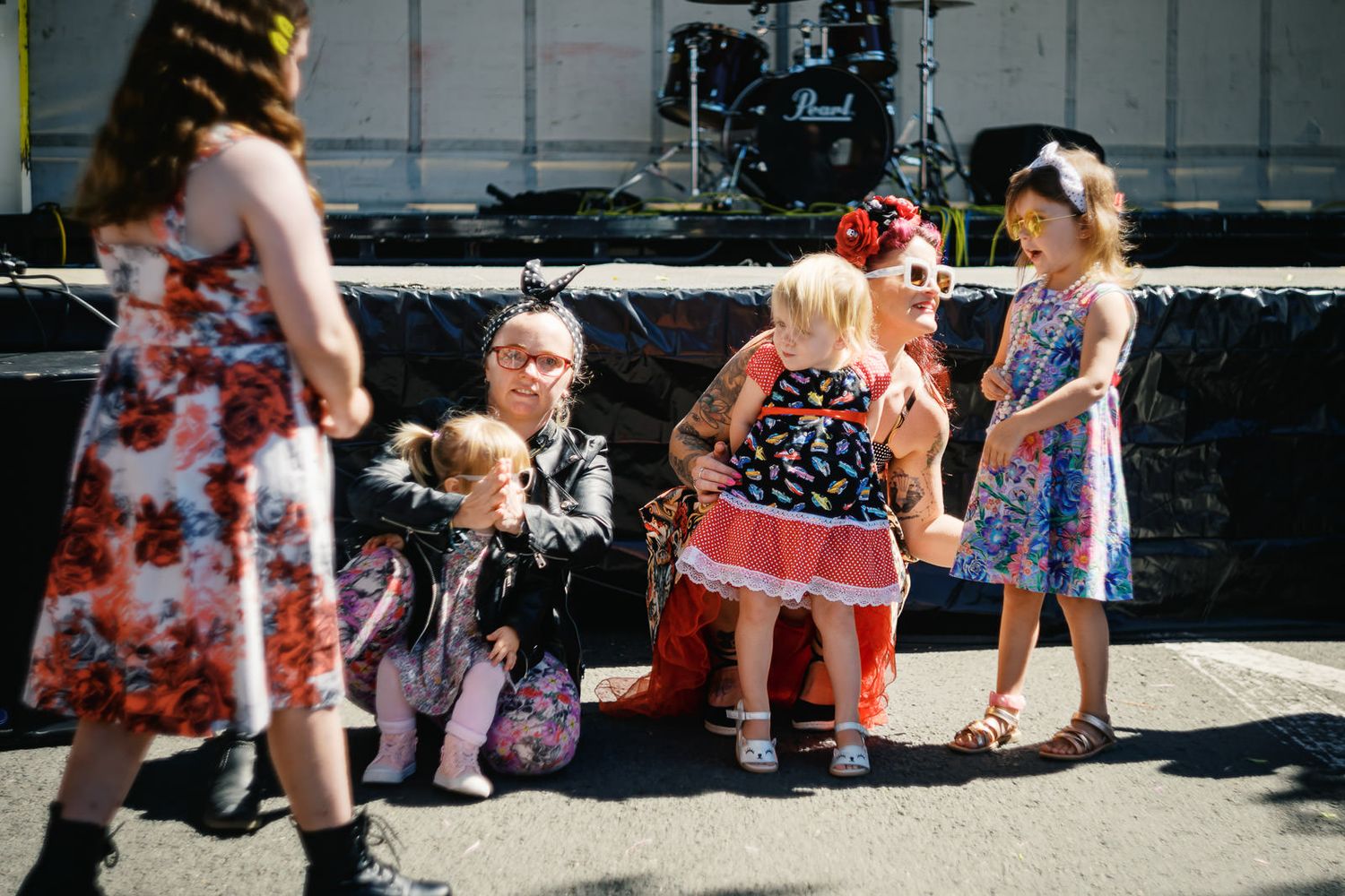 Adults and children in colorful floral dresses gather near an outdoor stage at a sunny festival event.
