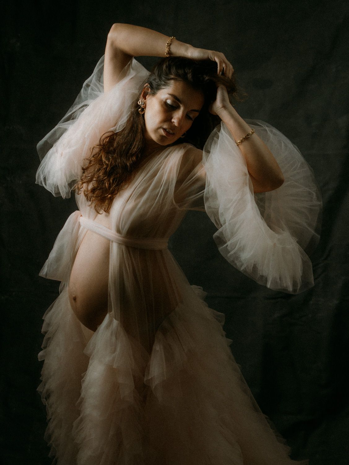 Pregnant woman in sheer white tulle gown poses gracefully against dark backdrop in elegant maternity portrait.