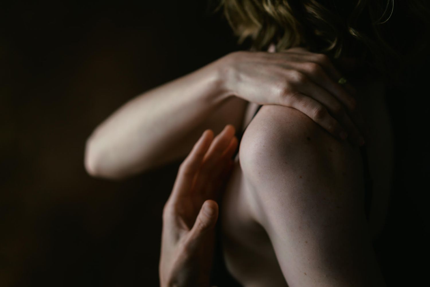 Intimate portrait of a person's bare shoulder and neck with hand gently touching skin in soft, dramatic lighting.