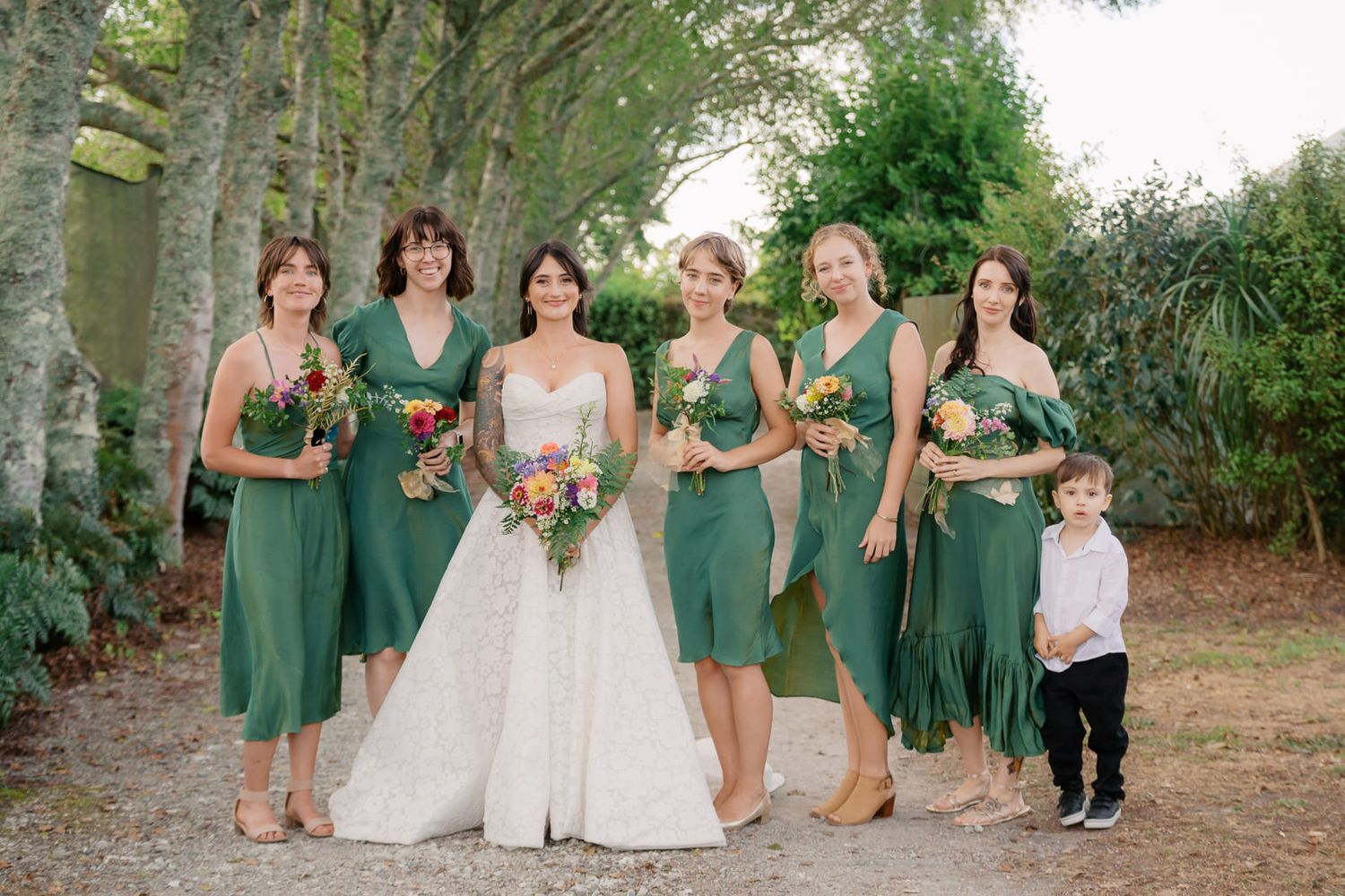 Bride in white gown with bridesmaids in green dresses holding colorful bouquets outdoors at a wedding.