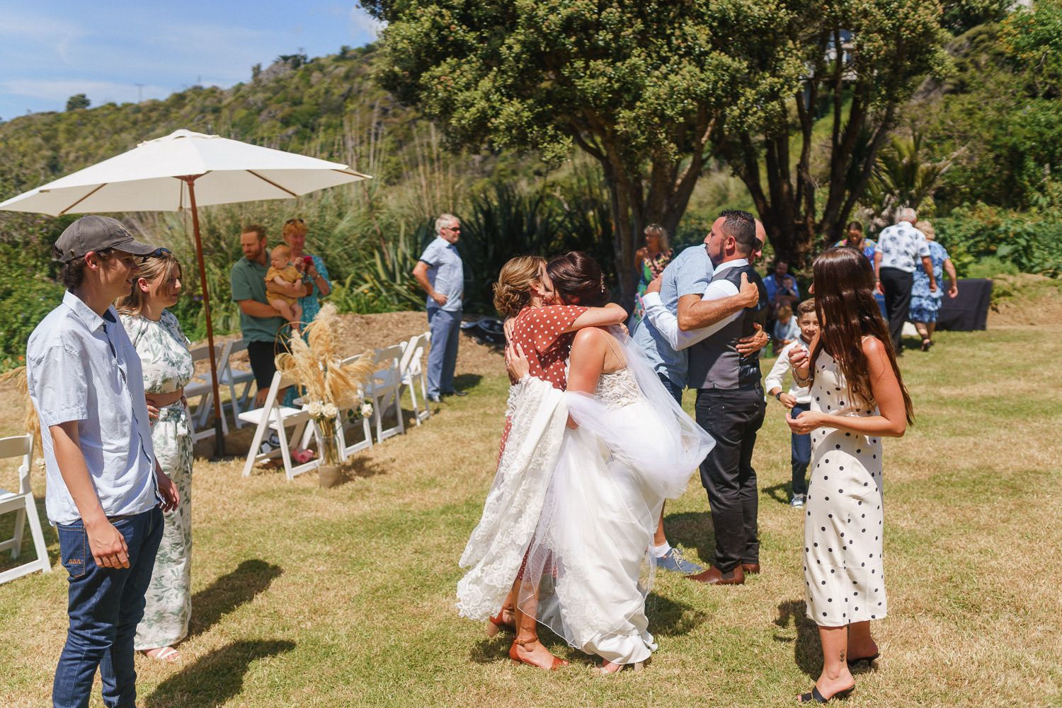 Bride in white dress hugging guests at outdoor wedding reception on sunny green lawn.