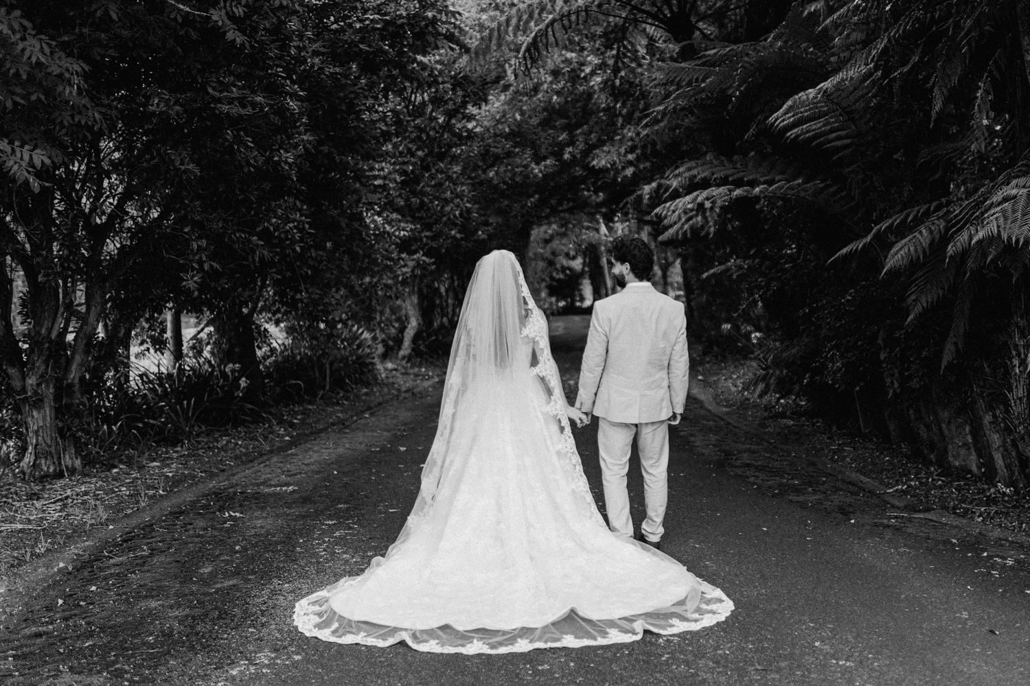 Black and white photo of bride and groom walking hand in hand down a tree-lined path, bride wearing long veil.
