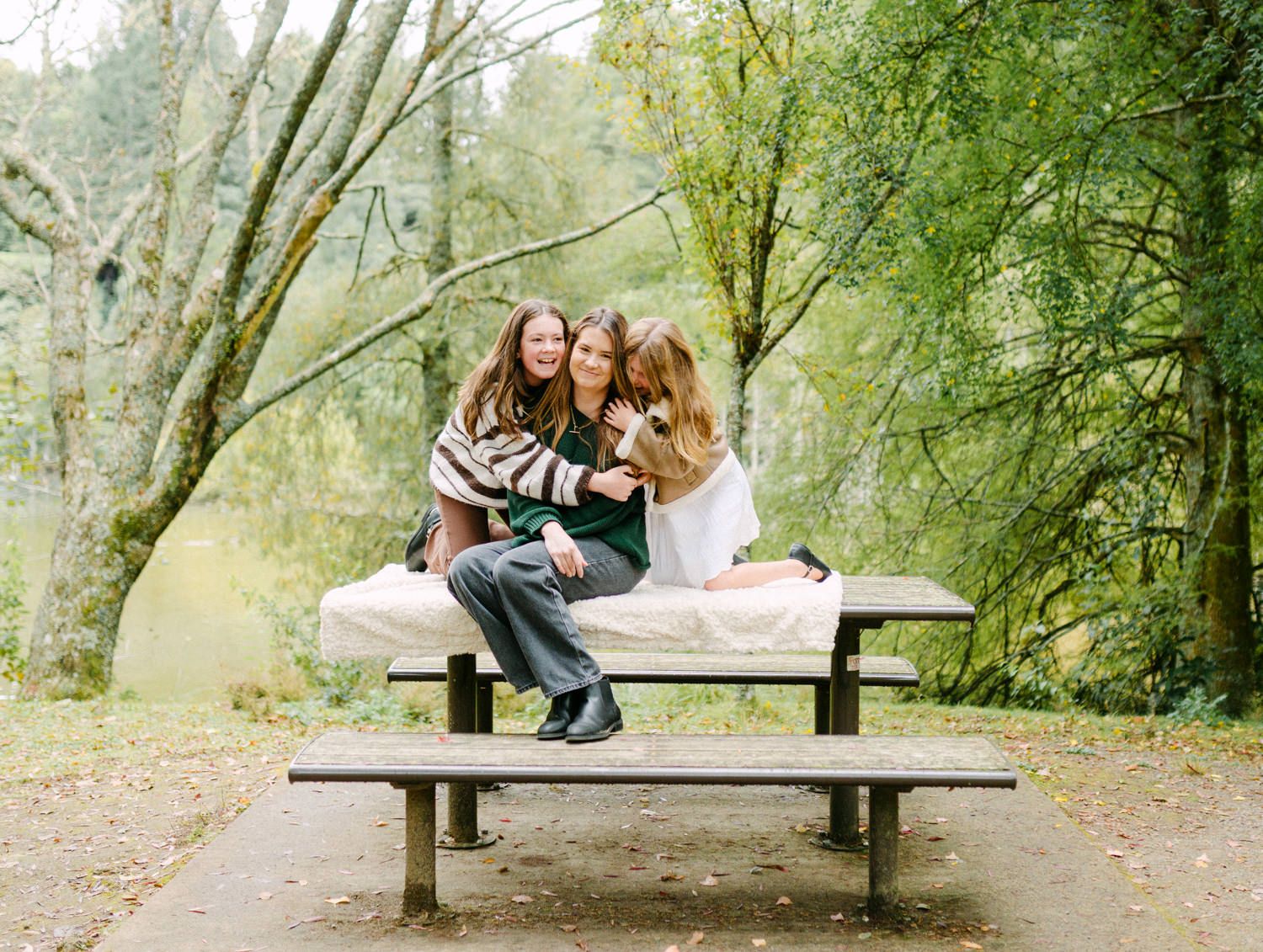 Three girls smiling and hugging on a picnic table surrounded by lush green trees in a park setting.