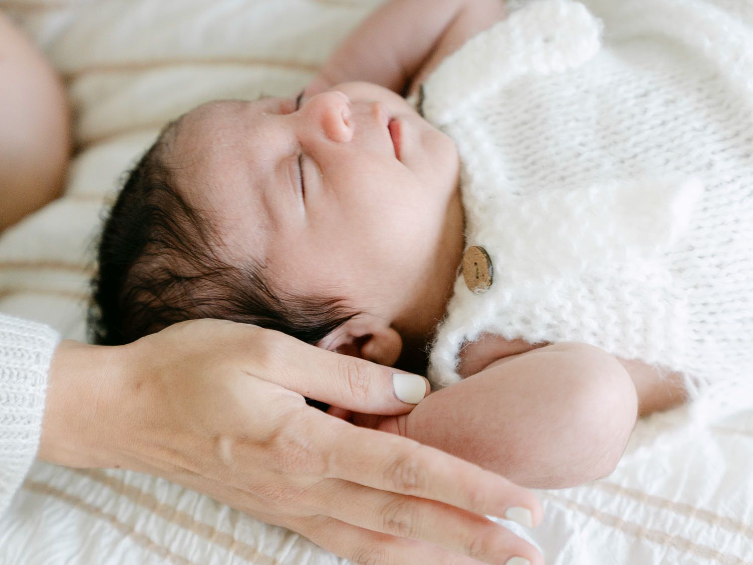 Newborn baby sleeping peacefully on white blanket with mother's hand gently supporting head.