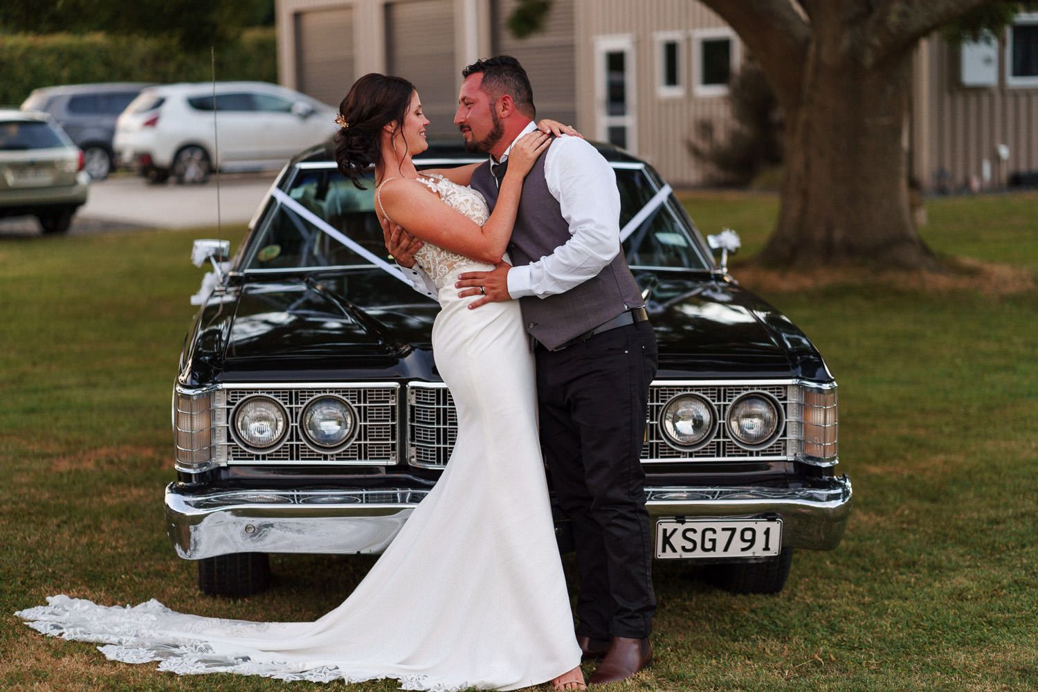 Bride and groom share a romantic embrace in front of a classic black vintage car decorated with white ribbons at their wedding.