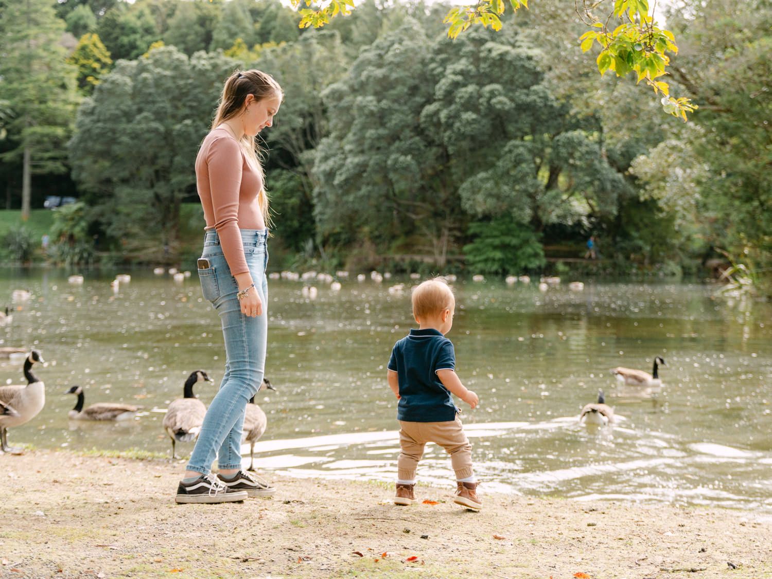 A mother and toddler feeding geese and ducks by a peaceful park pond on a sunny day.