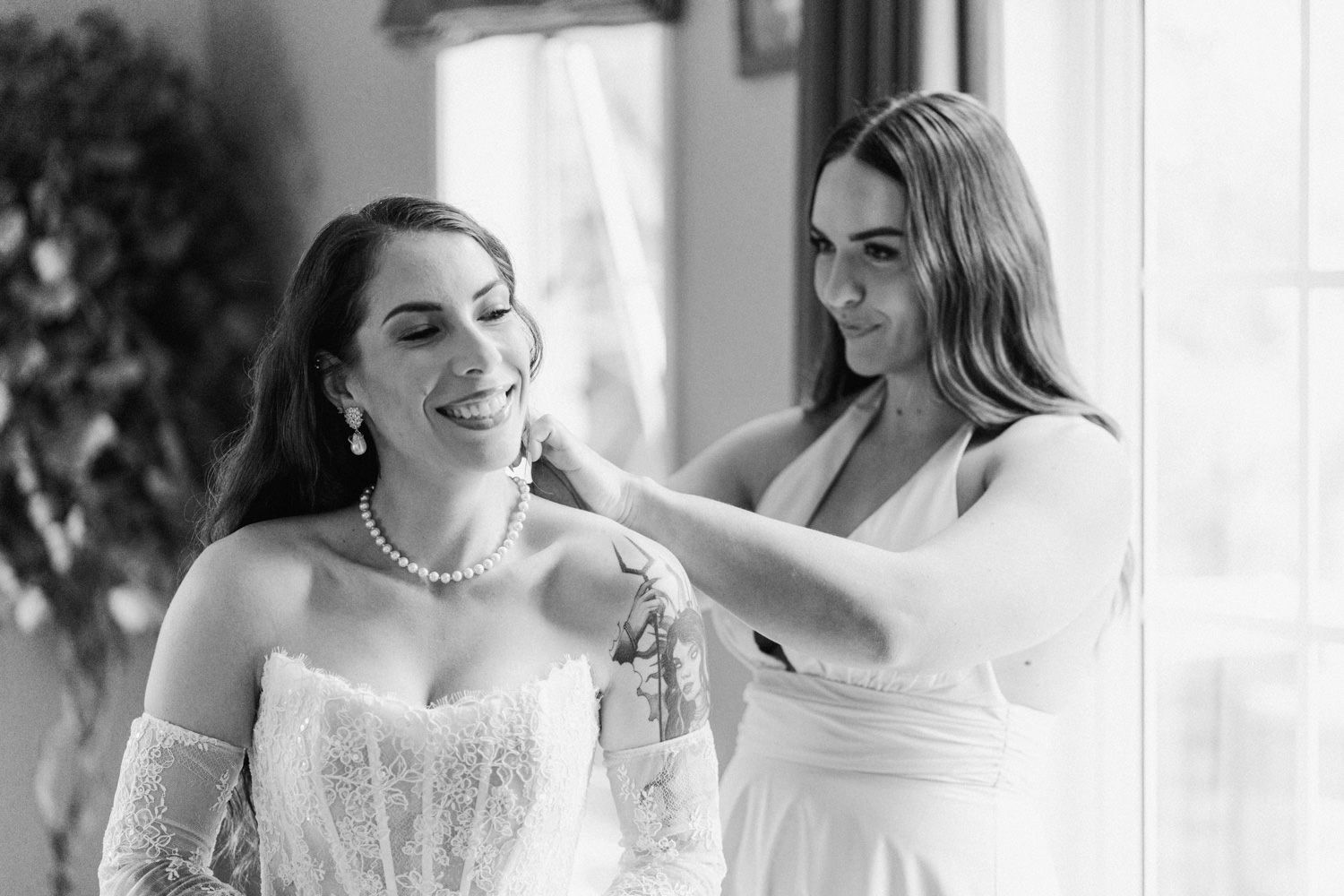 Bridesmaid helping bride with necklace while getting ready in white dresses, black and white wedding photo.