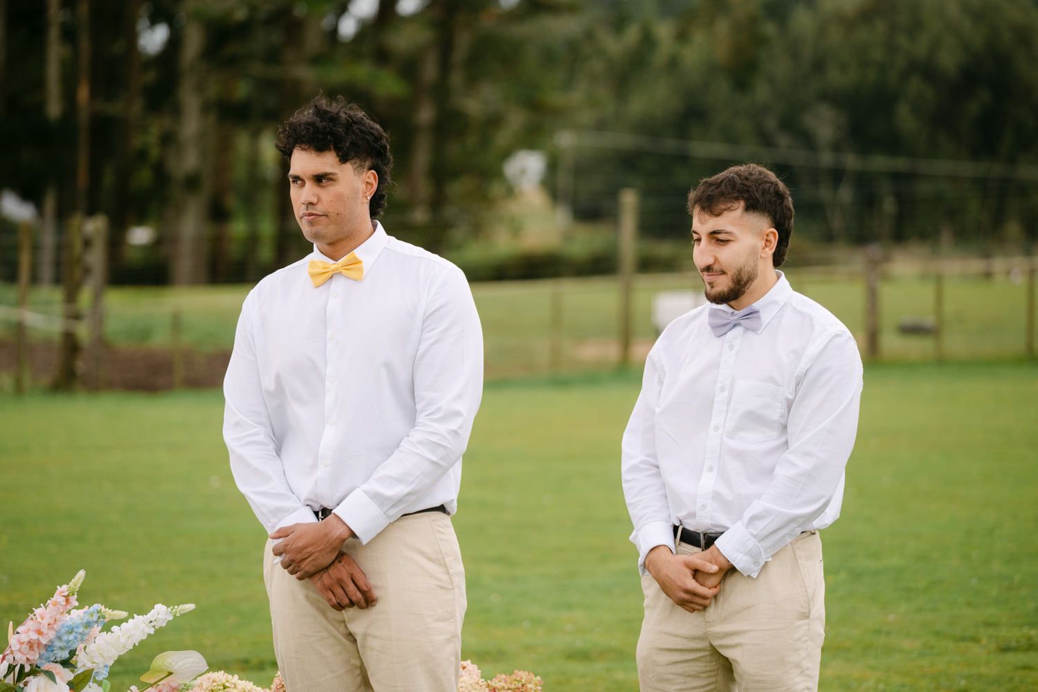 Two groomsmen in white shirts and bow ties standing outdoors at a wedding ceremony on a green lawn.