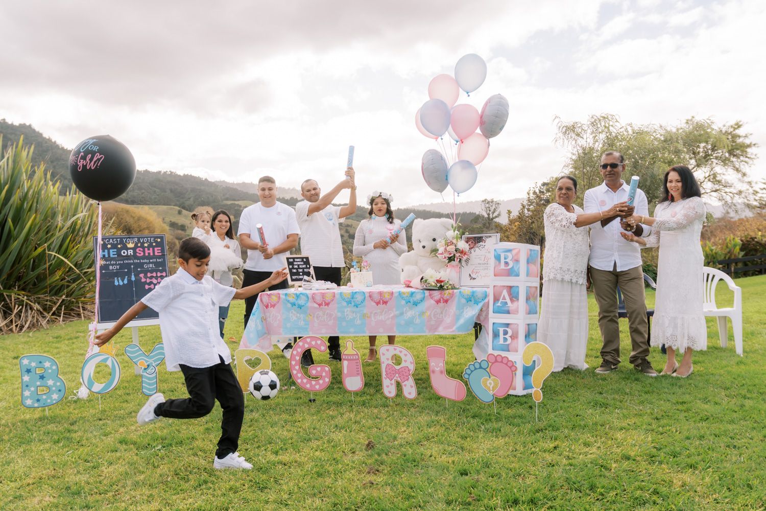 Gender reveal party with family holding BOY or GIRL signs, balloons, and a black balloon on a green lawn.