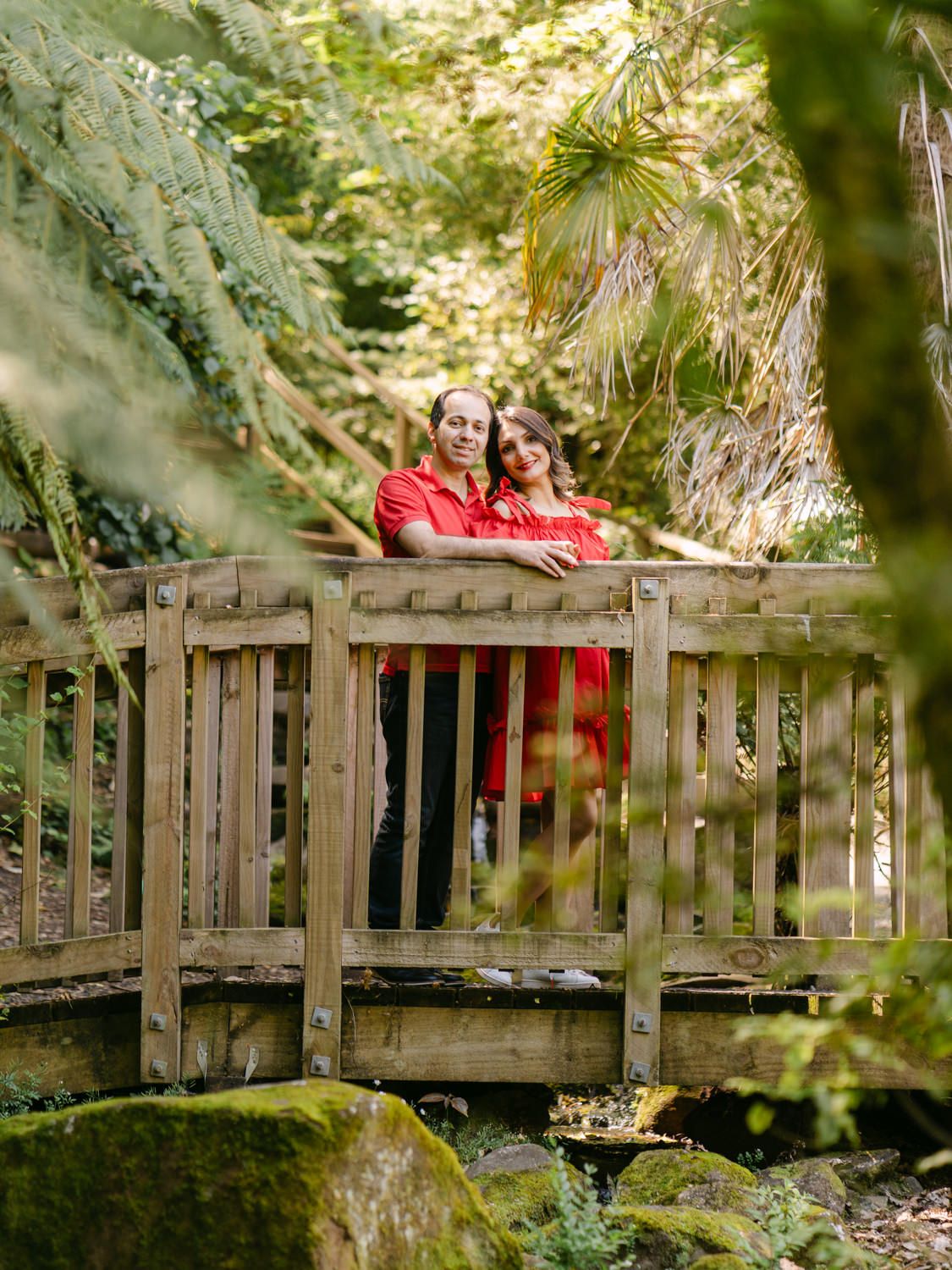 Couple in red outfits embracing on a wooden bridge surrounded by lush green forest foliage.