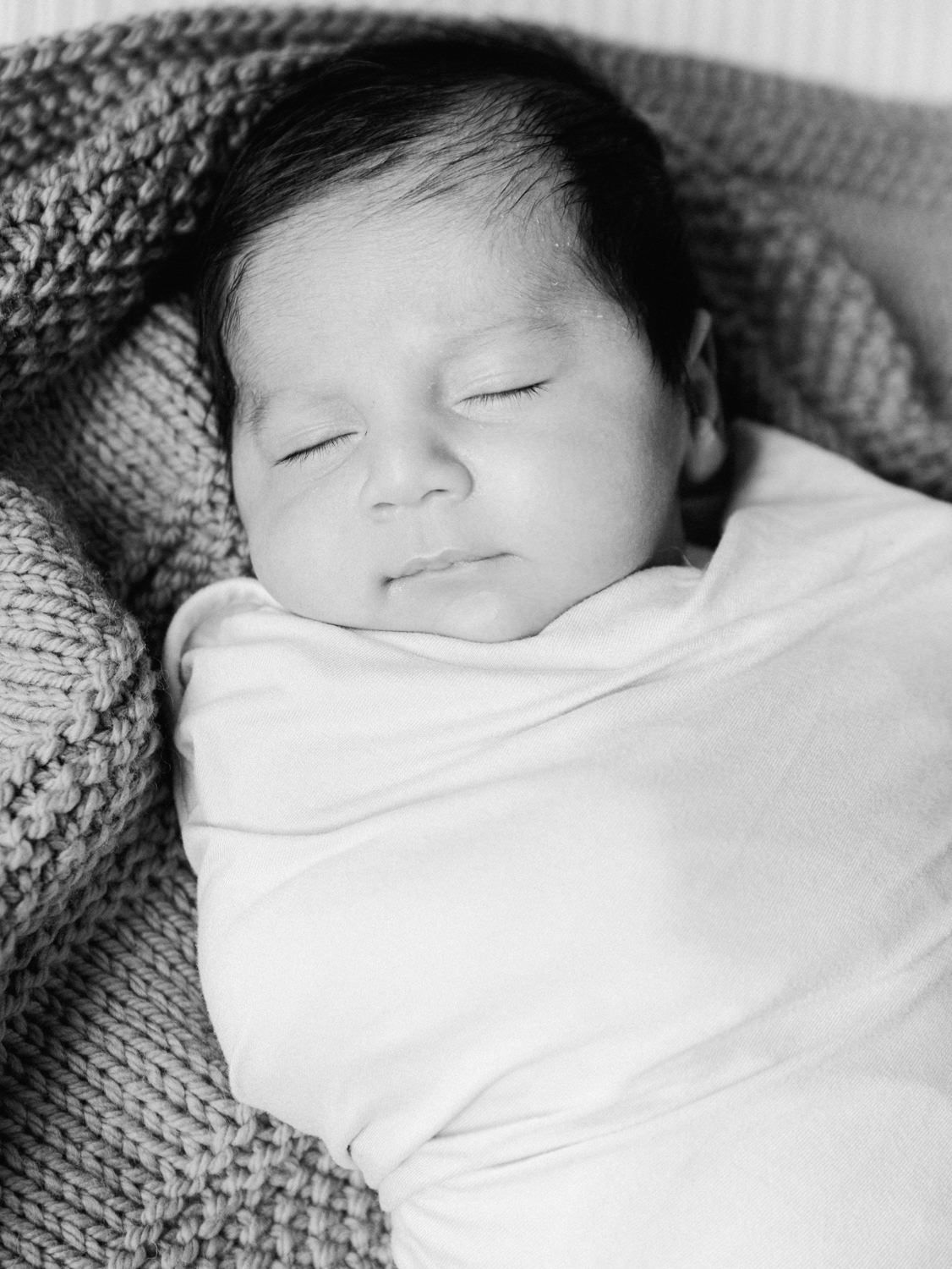 Close-up black and white portrait of a peacefully sleeping swaddled newborn baby with dark hair.