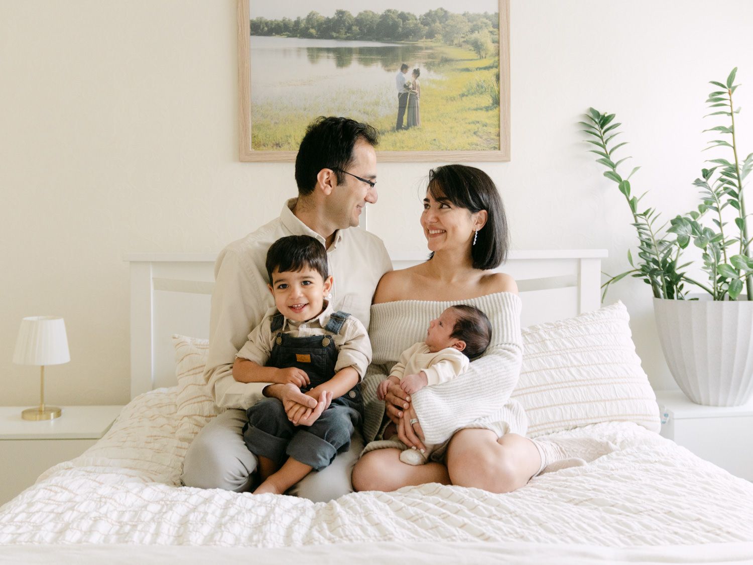 Happy family of four with toddler and newborn sitting together on a white bed, parents smiling at each other.