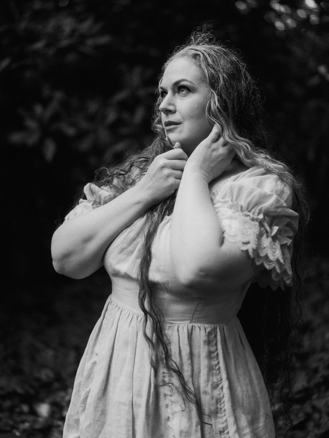 Black and white portrait of a woman in a vintage dress with long wavy hair, posing outdoors among lush greenery.