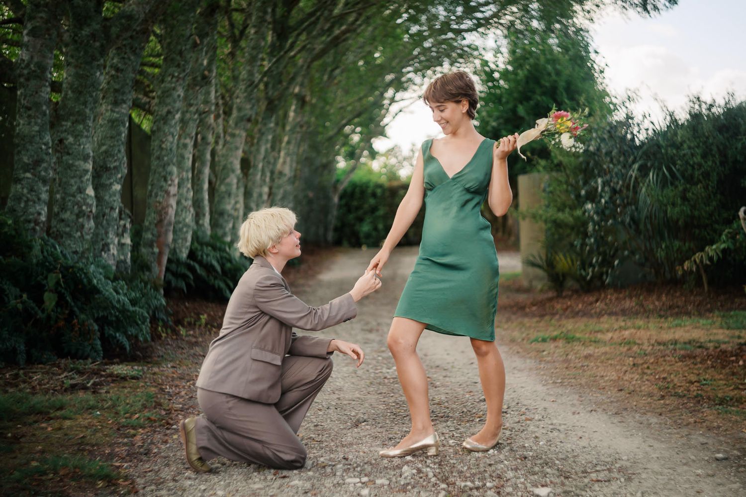 Two women in elegant dresses hold hands on a tree-lined gravel path, one kneeling gracefully outdoors.