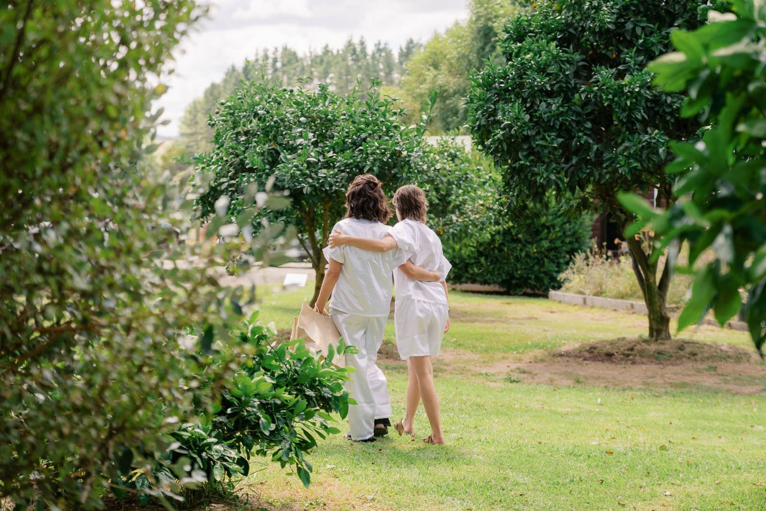 Two people in white outfits walk arm in arm through a lush green garden with trees and shrubs.