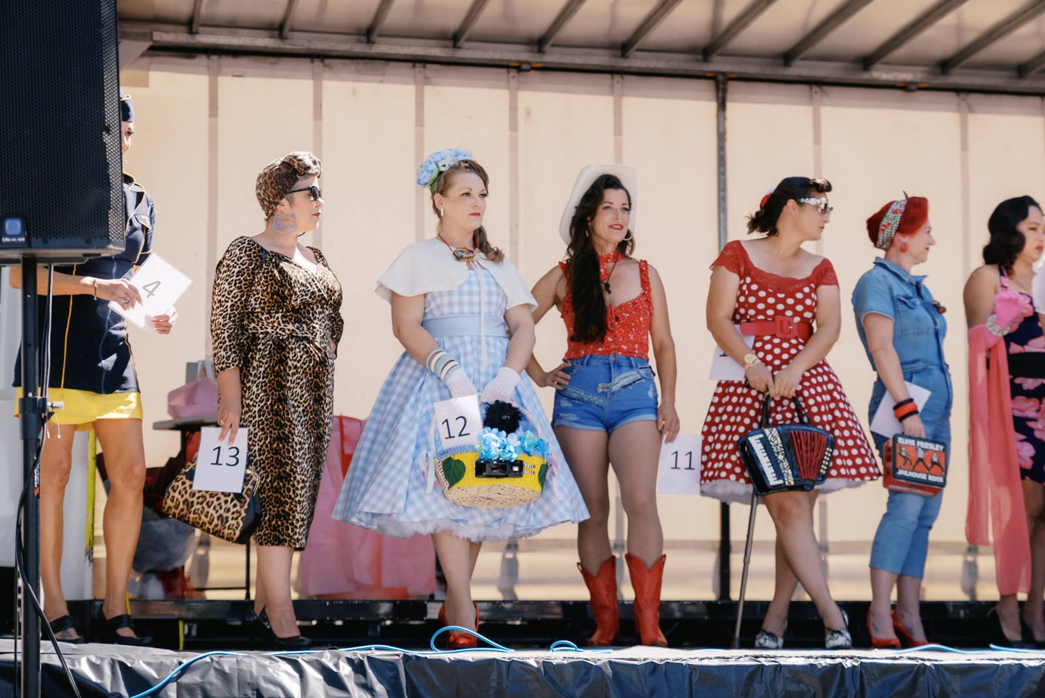 Women in vintage pin-up costumes compete in a retro costume contest on an outdoor stage at a fair.