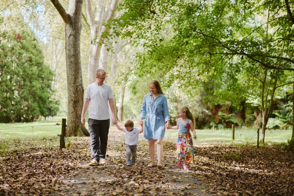 Family of four walking hand-in-hand on a leaf-covered path through a lush green park on a sunny day.