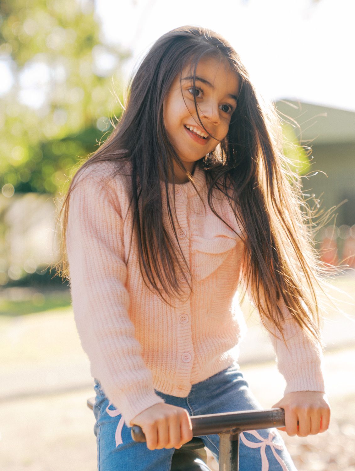 A smiling young girl with long dark hair wearing a pink cardigan and jeans, leaning on a railing outdoors.