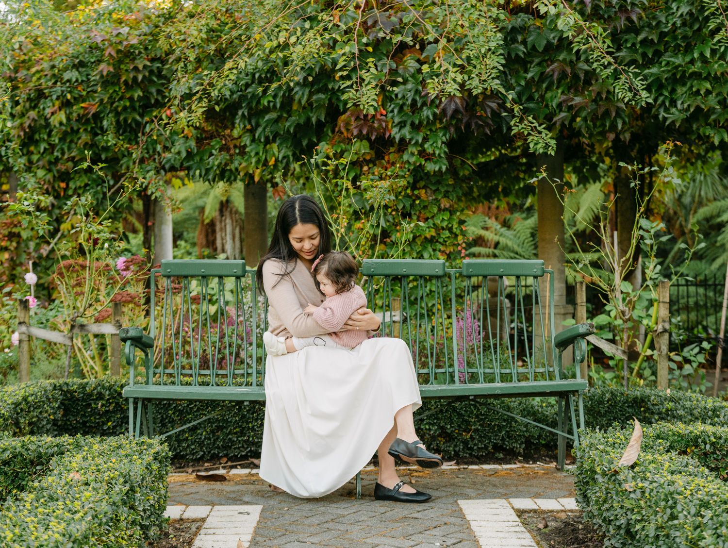 Mother cradling her baby on a green garden bench surrounded by lush greenery and flowering plants.