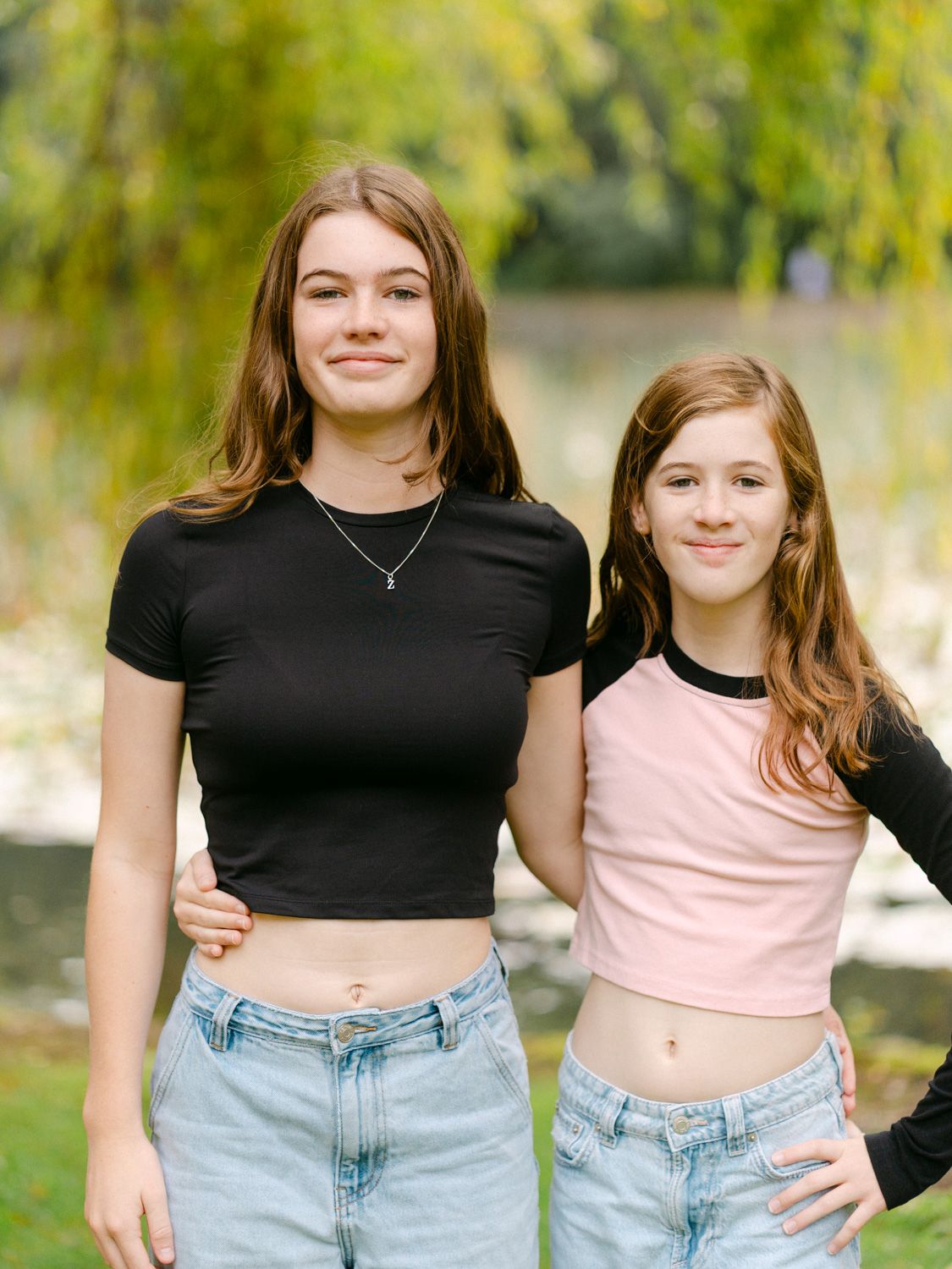 Two teenage girls smiling outdoors in casual crop tops and jeans with a green tree and pond in the background.