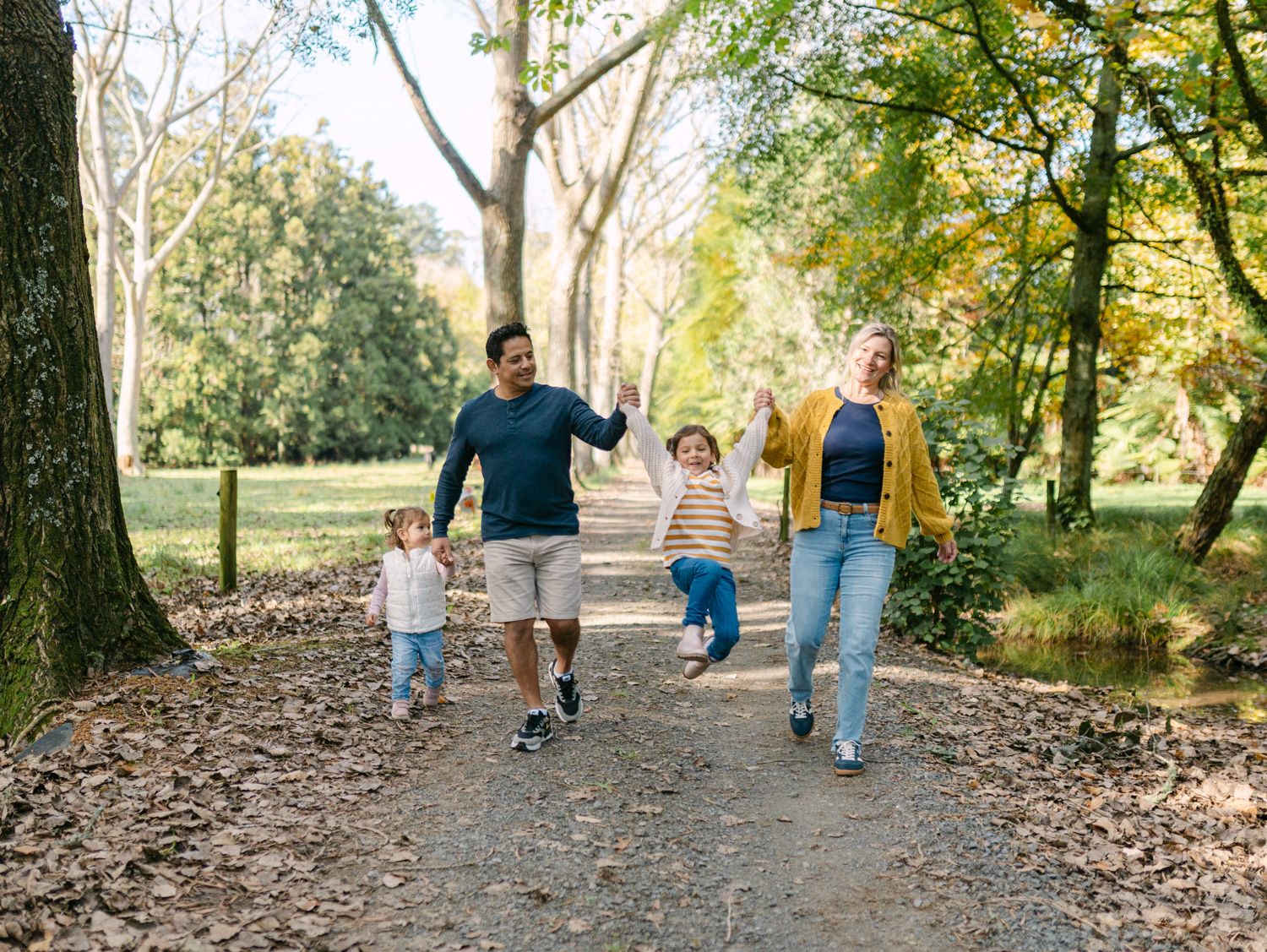 Family of four walking on a scenic park path surrounded by autumn trees, parents swinging children joyfully.