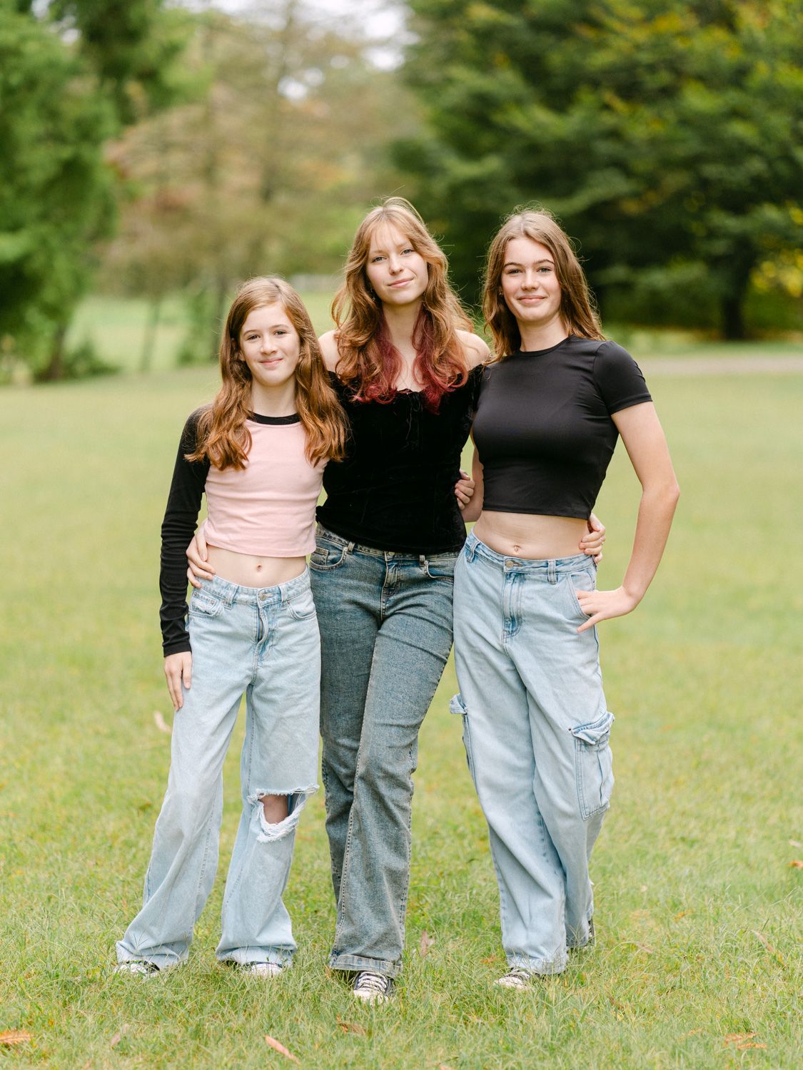 Three teenage girls with auburn hair pose together smiling outdoors in a green park, wearing jeans and crop tops.