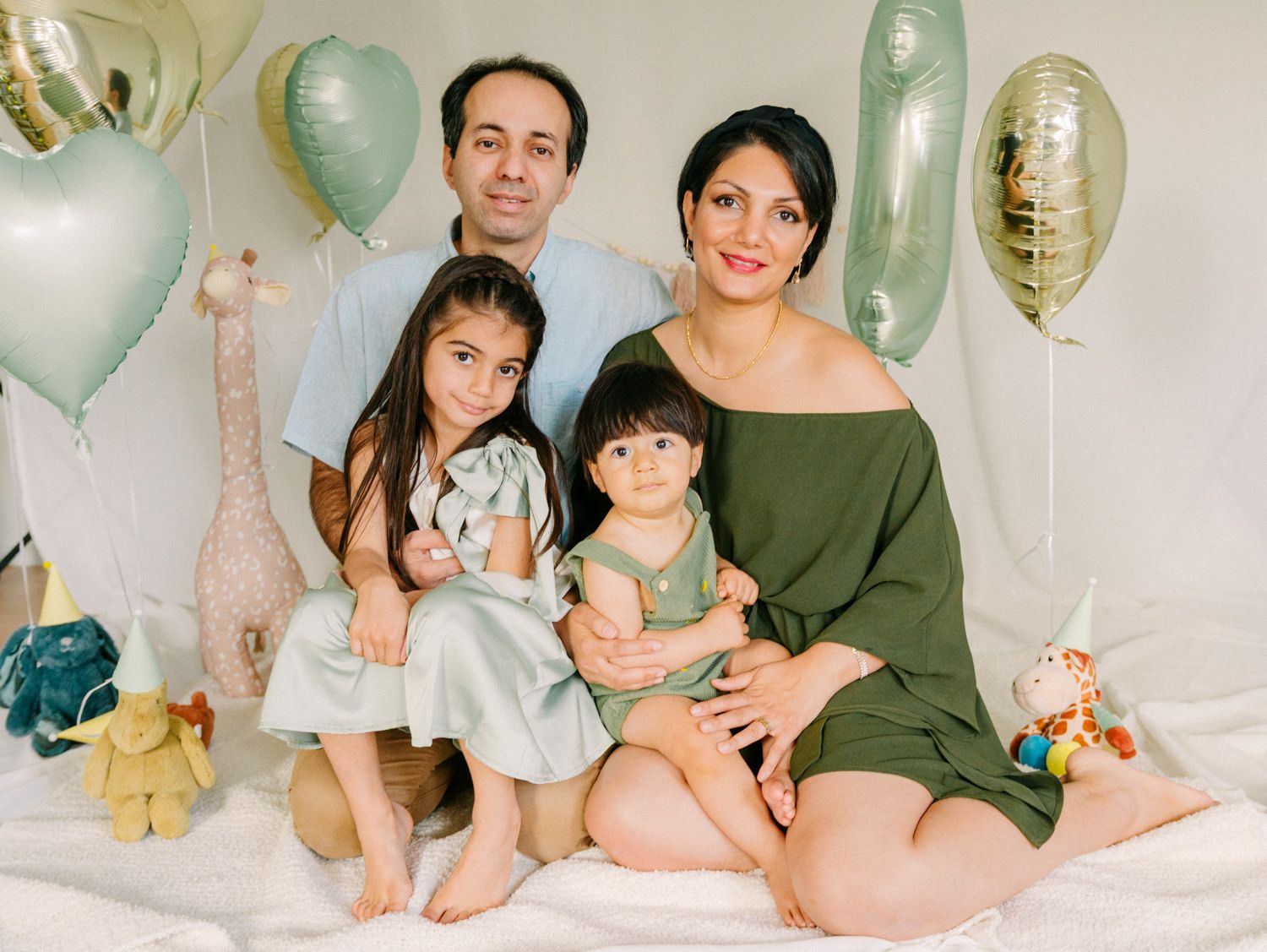 Family of four posing with pastel balloons and stuffed animals during a cheerful indoor photo session.