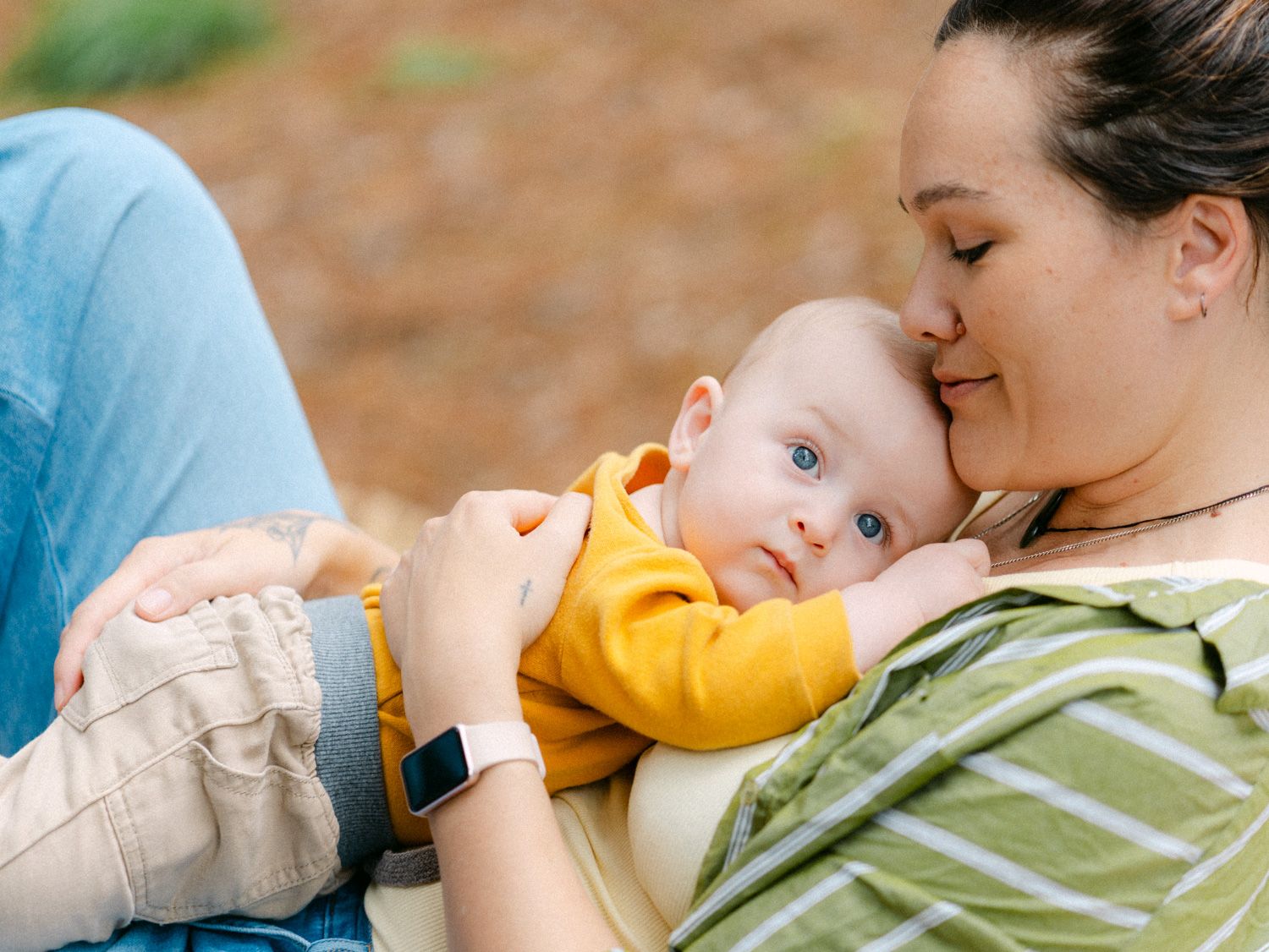 A mother tenderly cuddles her blue-eyed baby outdoors, both dressed casually in a warm, natural setting.
