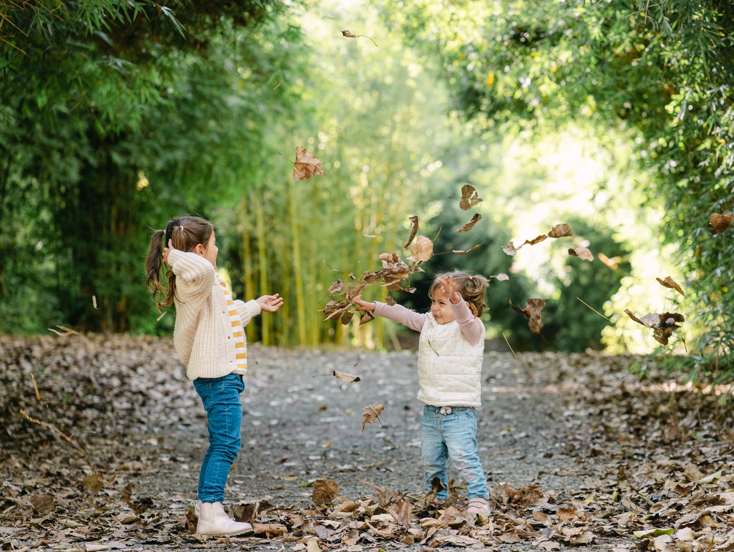 Two children joyfully tossing autumn leaves in a tree-lined path surrounded by lush green bamboo.