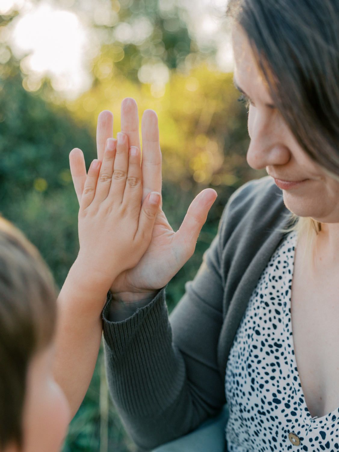 A mother and child pressing their hands together palm-to-palm outdoors in warm golden sunlight.