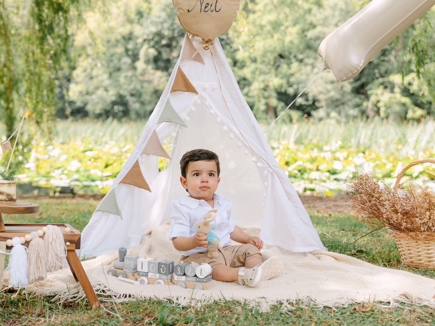 Baby boy sits on blanket before white teepee tent outdoors for first birthday photo shoot in park.