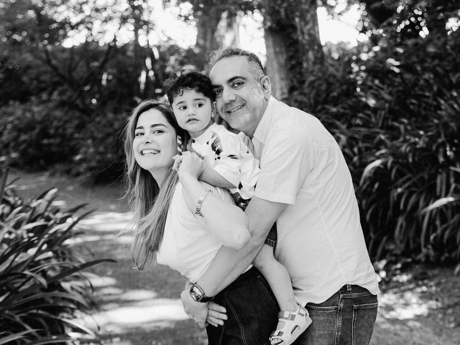Black and white family portrait of smiling parents holding young toddler outdoors among lush green trees.