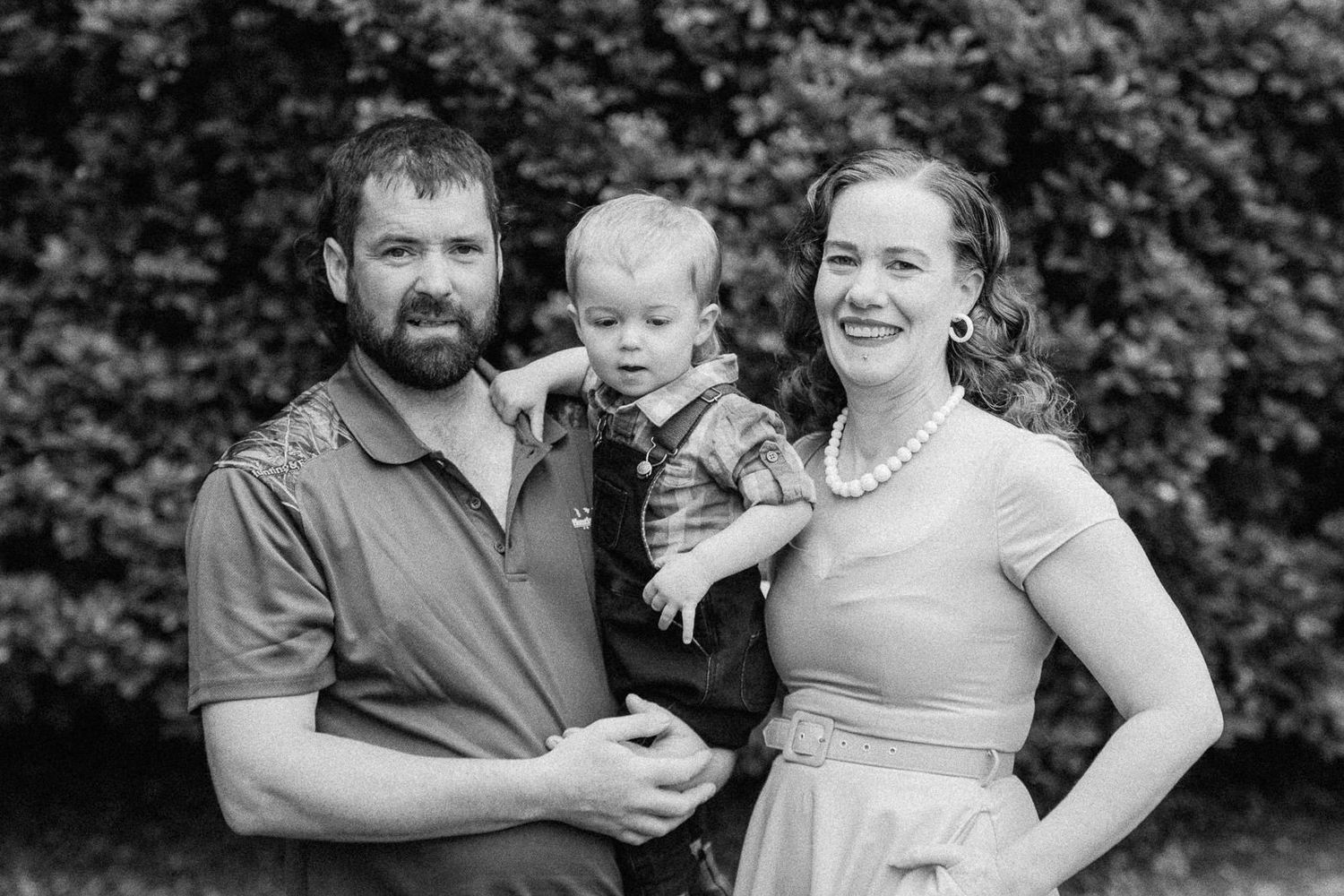 Black and white family portrait of bearded father, mother, and toddler son outdoors.
