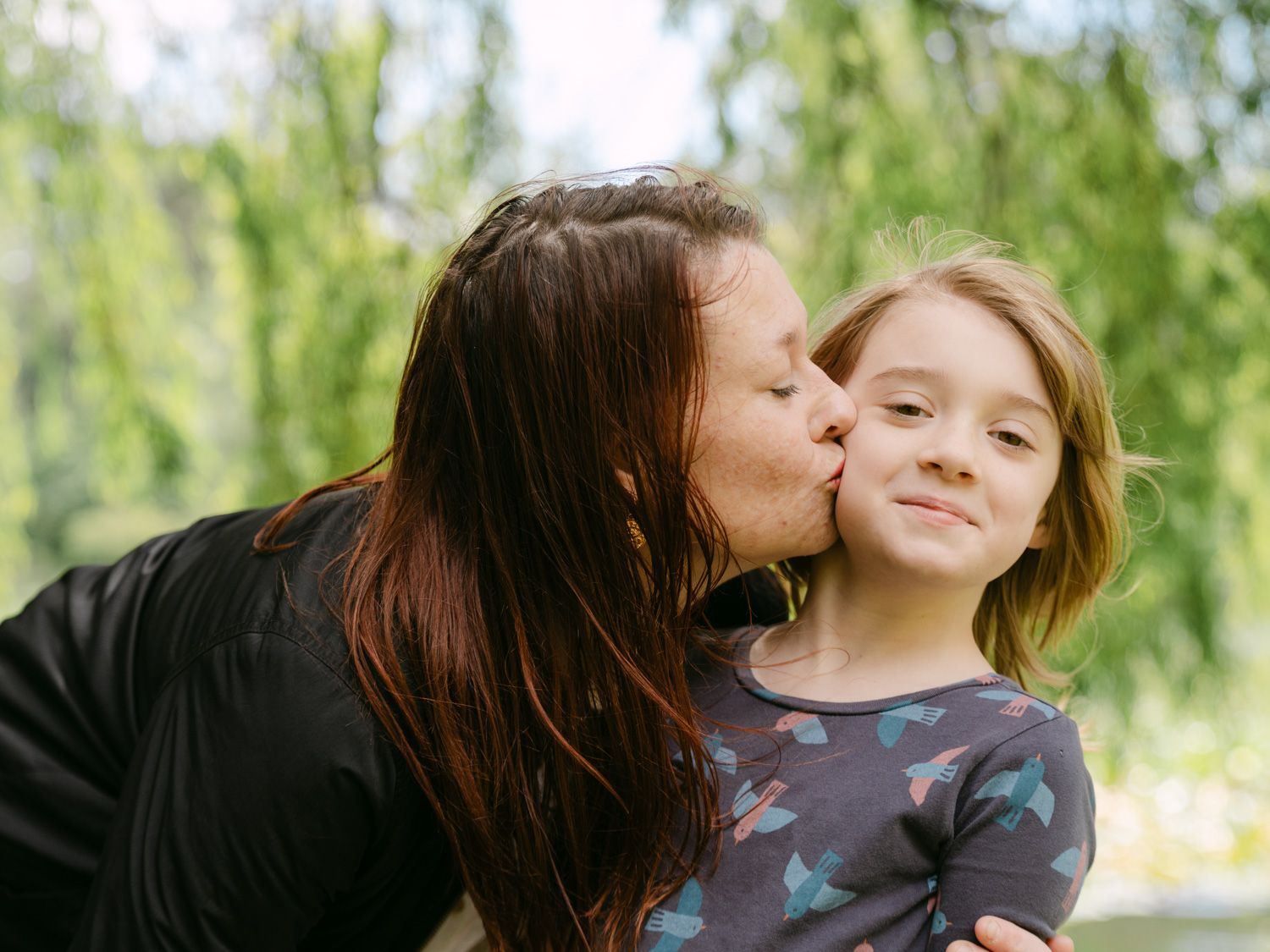 A mother lovingly kisses her young daughter on the cheek outdoors, with lush green trees in the background.