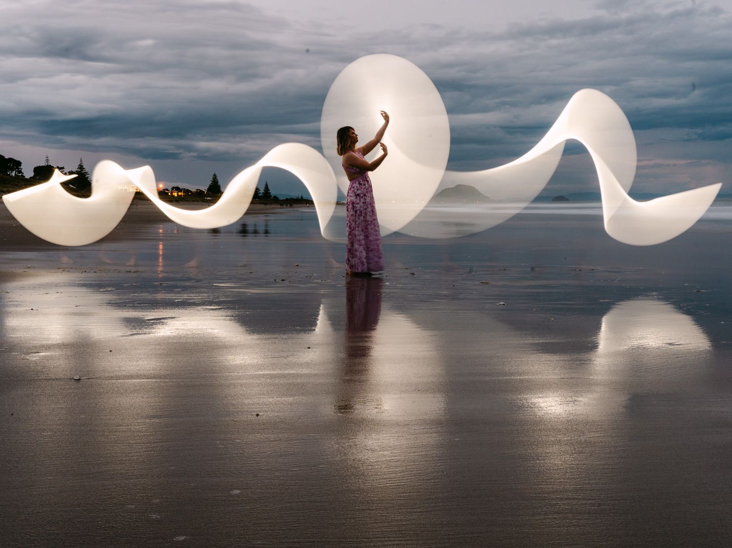 Woman in floral dress creating glowing light painting waves on a reflective beach at dusk.