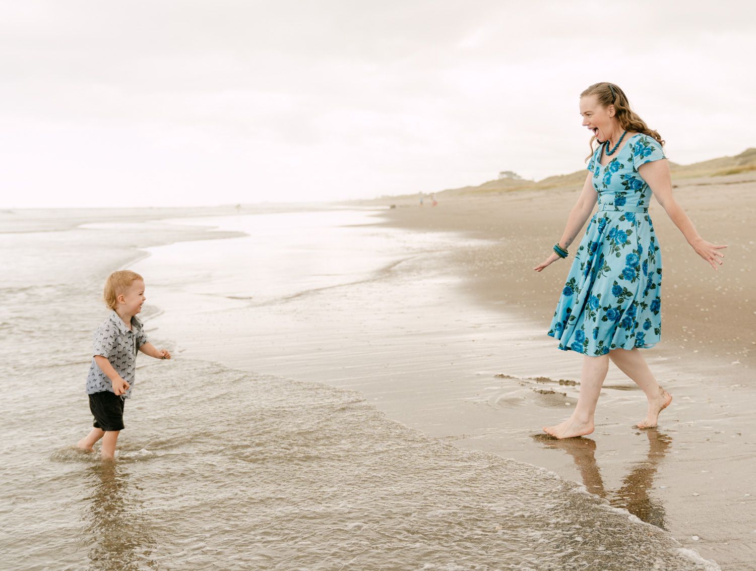 A woman in a blue floral dress plays with a toddler boy at the ocean's edge on a sandy beach.