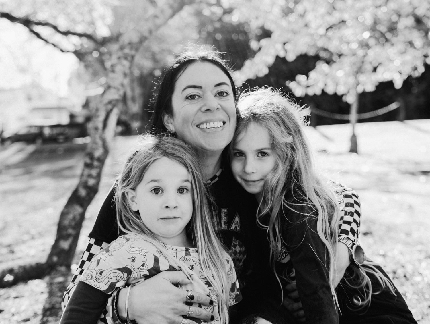 A smiling mother poses with her two daughters under a blossoming tree in a black-and-white park portrait.