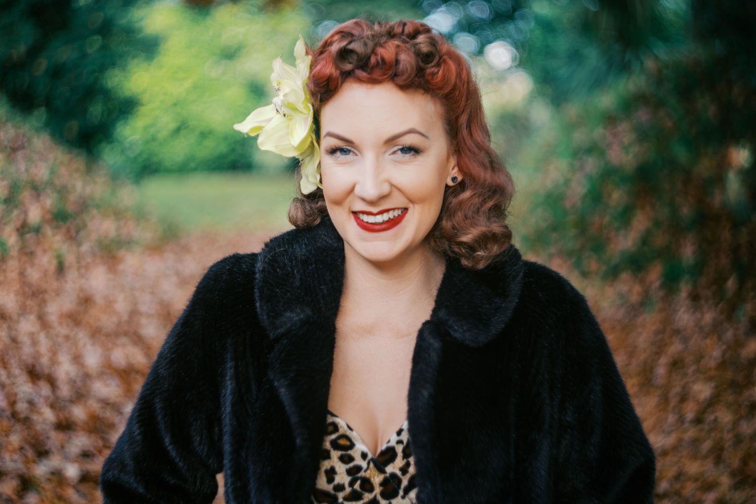 Smiling woman with red hair and flower accessory wearing a black fur coat and leopard print top outdoors.