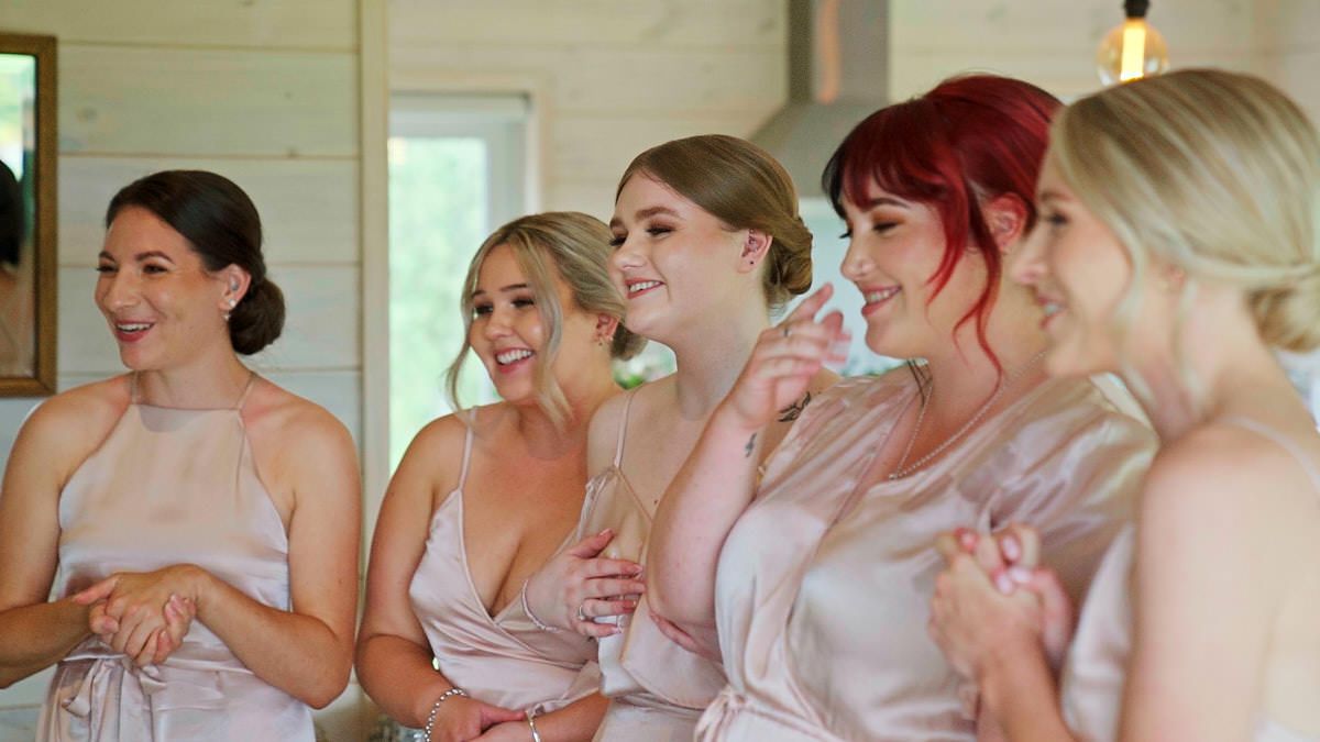 Five bridesmaids in blush satin dresses laughing and smiling together during wedding preparations indoors.