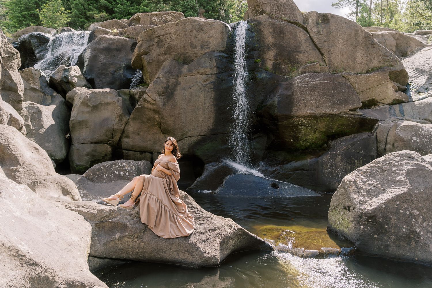 A woman in a flowing golden gown poses gracefully on rocks beside a cascading waterfall in a serene forest setting.
