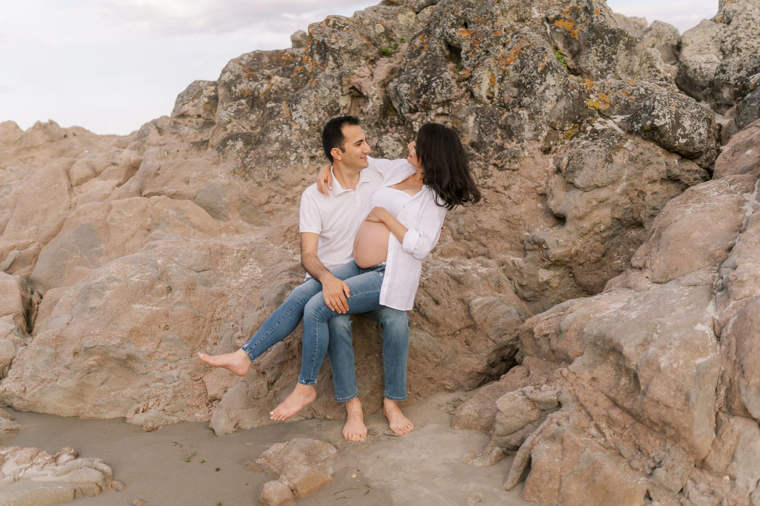 A couple sits barefoot on rocky beach boulders, smiling at each other in white shirts and jeans.