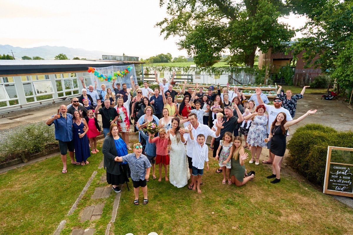 Large wedding group photo outdoors with guests waving and smiling in a garden setting near a building.