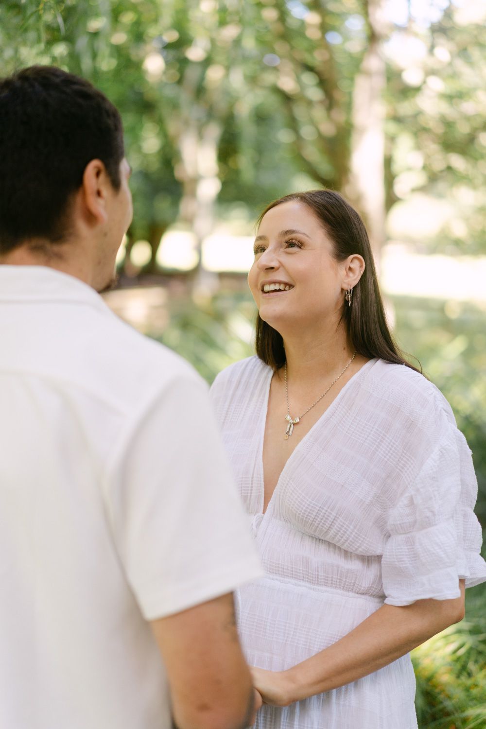 Smiling woman in white dress holding hands with man outdoors in lush green garden setting.