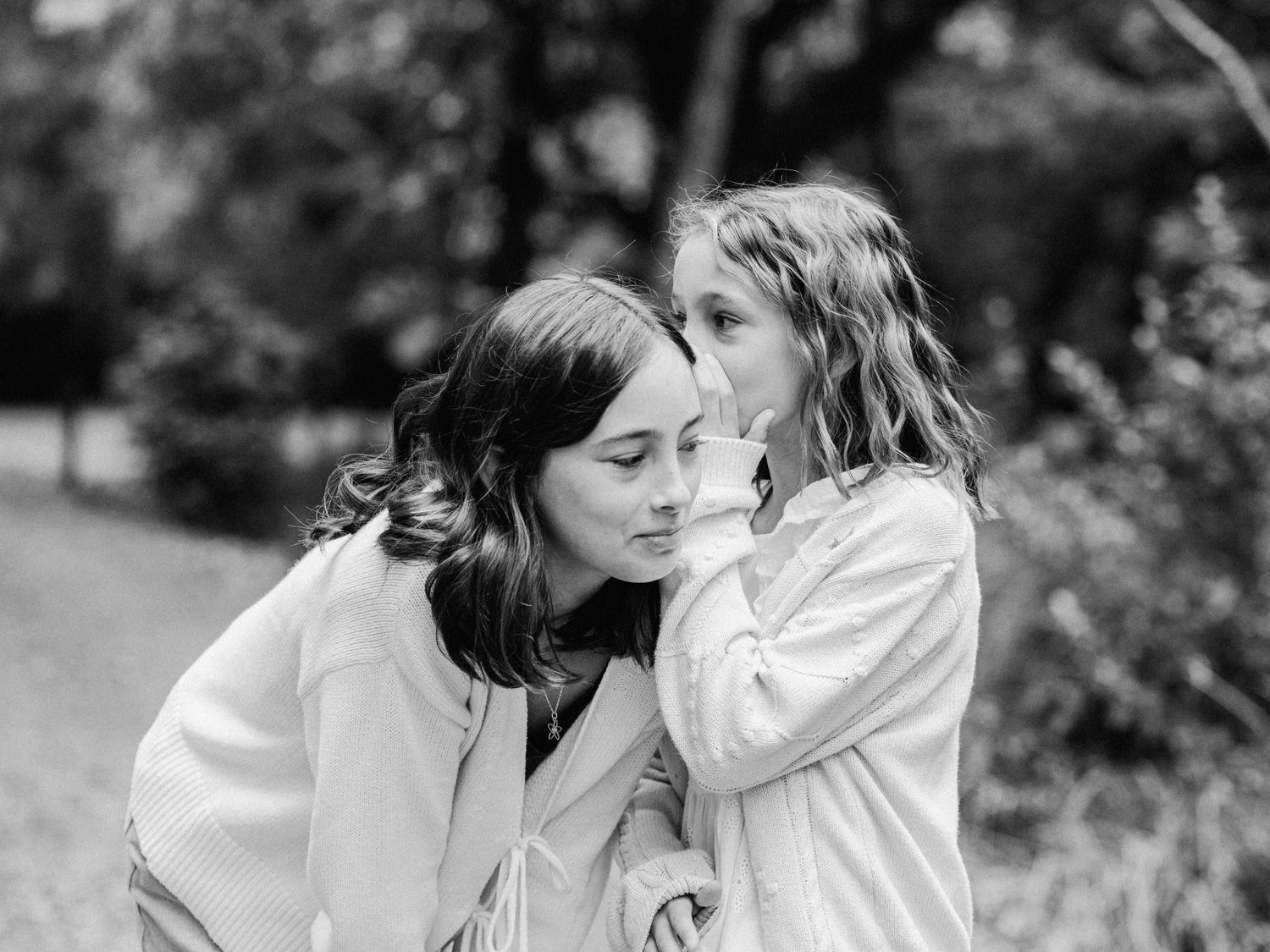 A young girl whispers a secret into a woman's ear outdoors in a black and white photo.