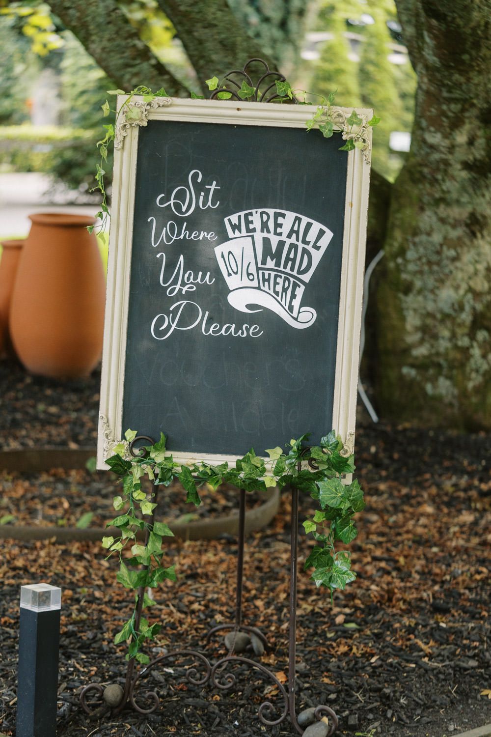 Chalkboard wedding sign on easel reads Sit Where You Please, We're All Mad Here, decorated with ivy.