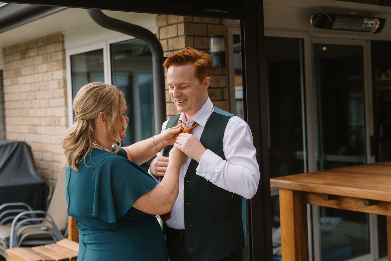 A woman in teal adjusts a boutonniere on a smiling redhead groom in a white shirt outdoors.